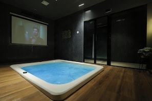 Habitació moderna amb un gran pantalla que mostra una persona, una banyera d'hidromassatge i una porta corredissa.