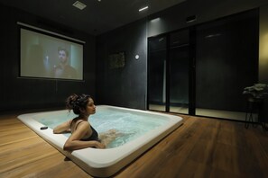 A person relaxing in a modern indoor hot tub with a large screen displaying a movie in the background. - Myotel - Andheri (Mumbai)