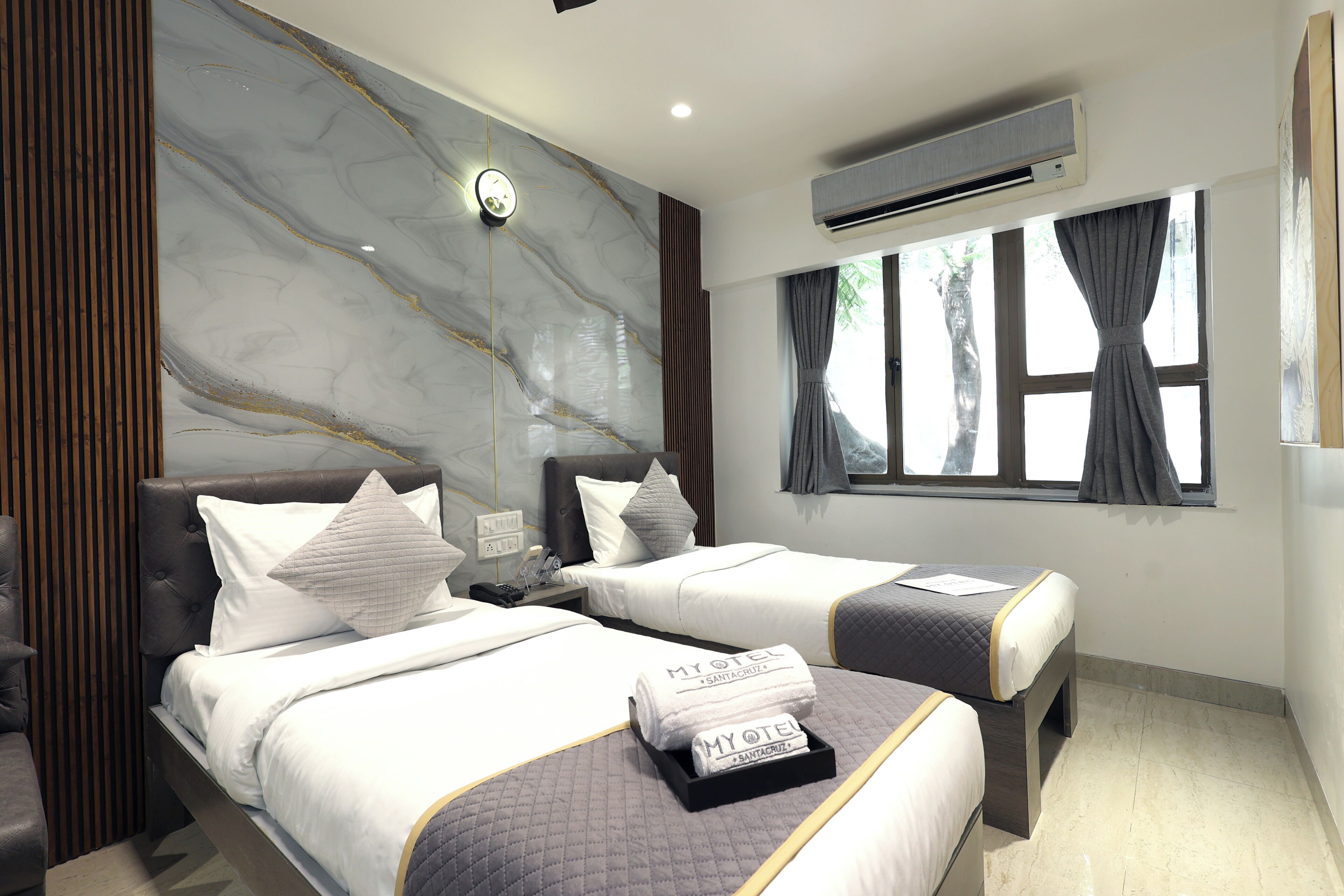 A hotel room with two beds, a marble wall, an air conditioner, and a window with curtains.