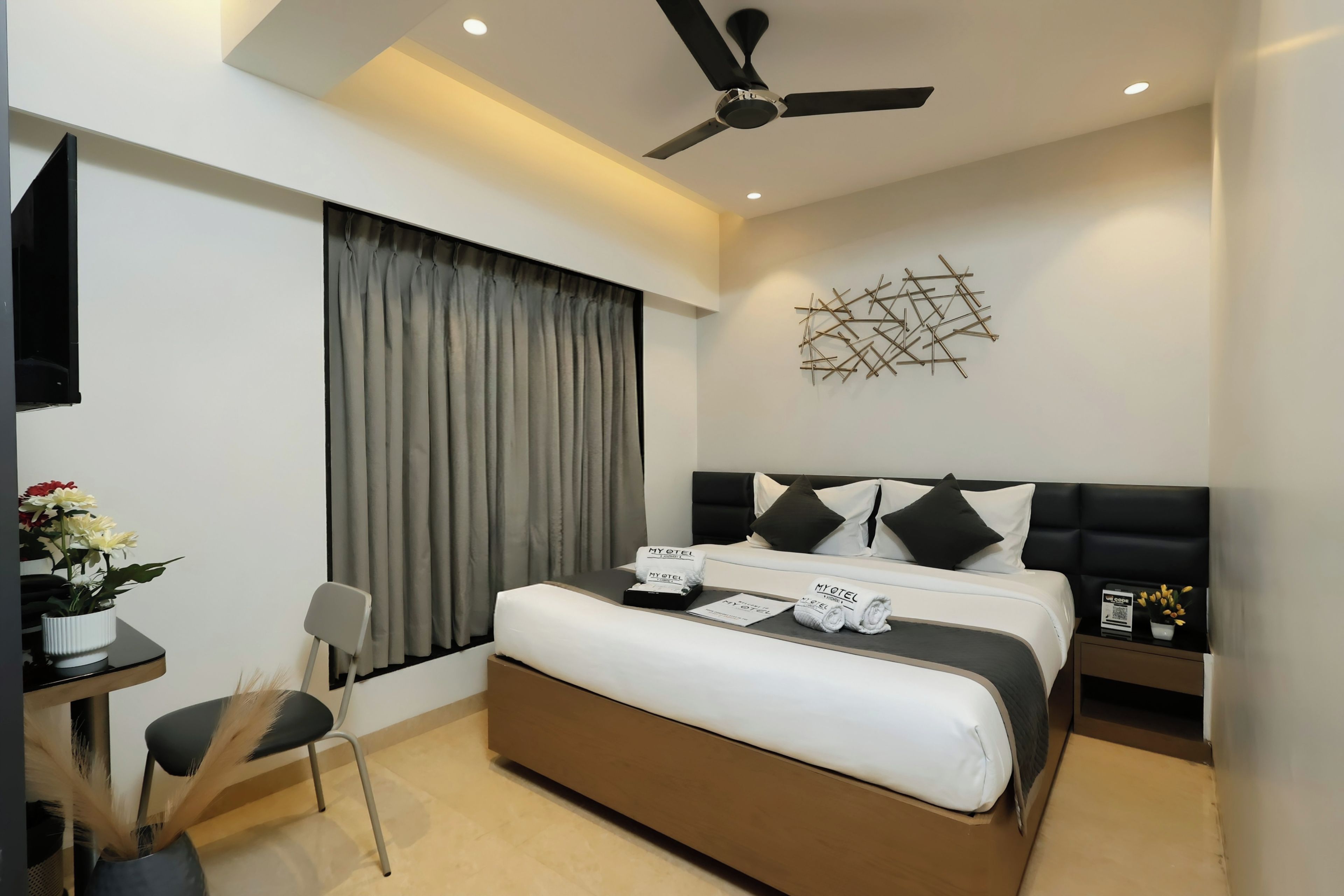 A modern hotel room with a bed, a ceiling fan, a wall-mounted abstract art piece, and a small table with a vase of flowers.