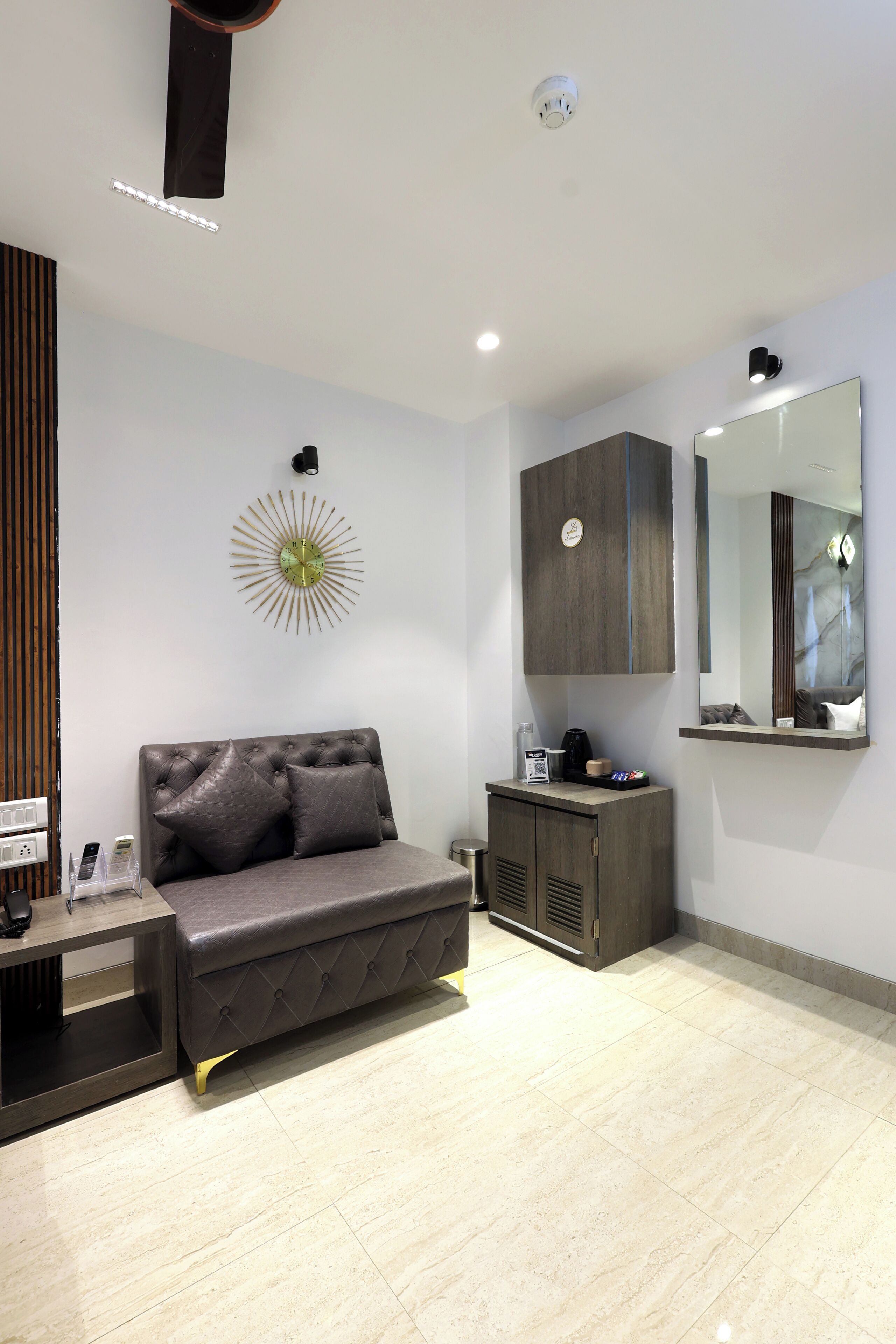 A modern bedroom with a grey sofa, a bedside table with a lamp, a small cabinet, and a wall-mounted mirror.