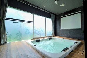 A modern spa room with a large hot tub, a sliding glass door leading to a garden, and a projector screen on the wall.