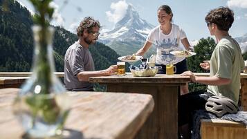 Ein Holztisch mit Essen und Getränken, Blick auf Berge und eine Person, die Speisen serviert.