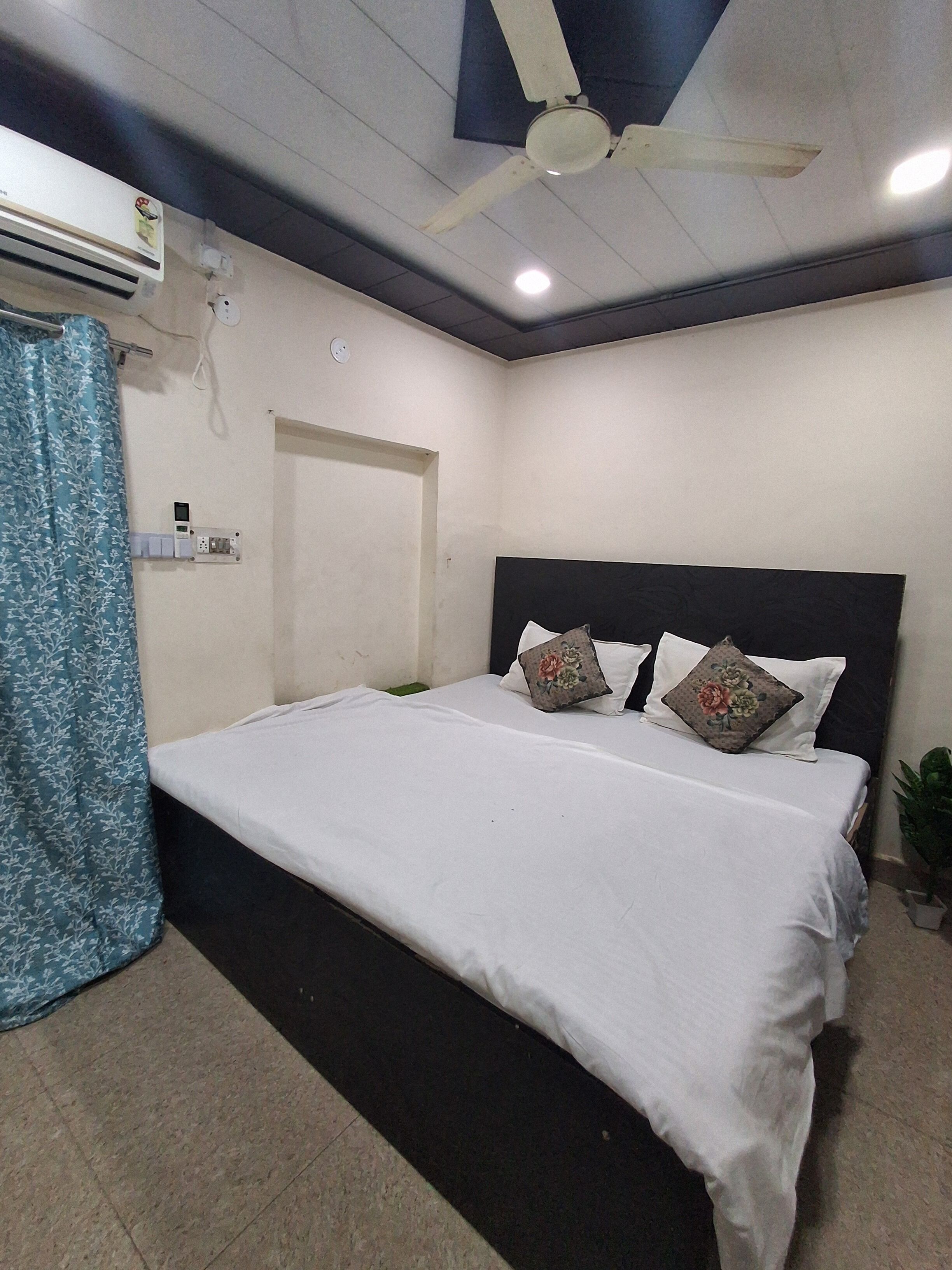 A bedroom with a bed, a ceiling fan, an air conditioning unit, a door, and a potted plant.