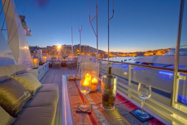 Terraza con sofá, velas, una botella y copas, con vista a un paisaje urbano al atardecer.