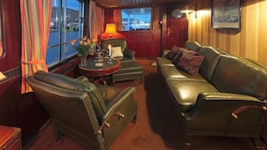 A cabin with leather furniture, a round table with a lamp, and a window with curtains. - Sanssouci Star - Exclusive Island Cruise (Flensburg)