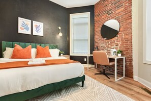 Ein modernes Schlafzimmer mit einem grünen gepolsterten Kopfteil, einem großen Bett mit orange-weißer Bettwäsche, einem Schreibtisch mit Stuhl und einem runden Spiegel an einer Ziegelwand.