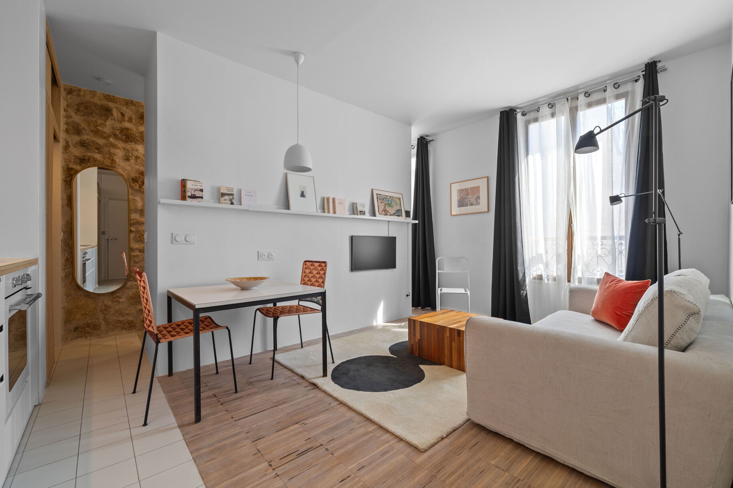 A modern living room with a sofa, a dining table, and chairs. There is a flat-screen TV mounted on the wall, a lamp, and a window with curtains.
