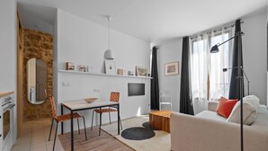 A modern living room with a sofa, a dining table, and chairs. There is a flat-screen TV mounted on the wall, a lamp, and a window with curtains. - Nid d'Ivresse, 1BR/1BA, 2 people (Paris)
