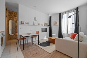 Ein stilvolles Wohnzimmer mit einer Couch, einem Esstisch und Stühlen. An der Wand ist ein Flachbildfernseher angebracht, ein Lampenschirm und ein Fenster mit Vorhängen.