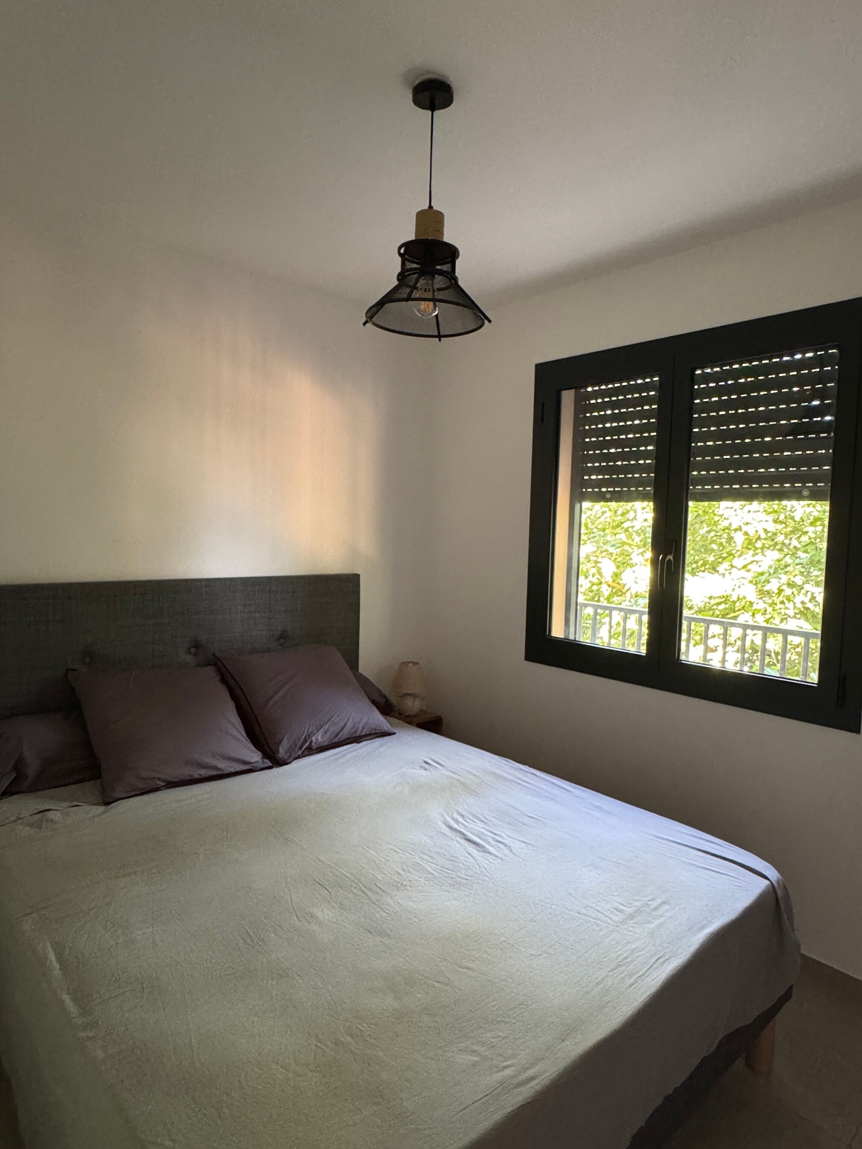 Ein Schlafzimmer mit einem Bett, einem Nachttisch, einer Lampe, einem Fenster mit Jalousien und einer Pendelleuchte.
