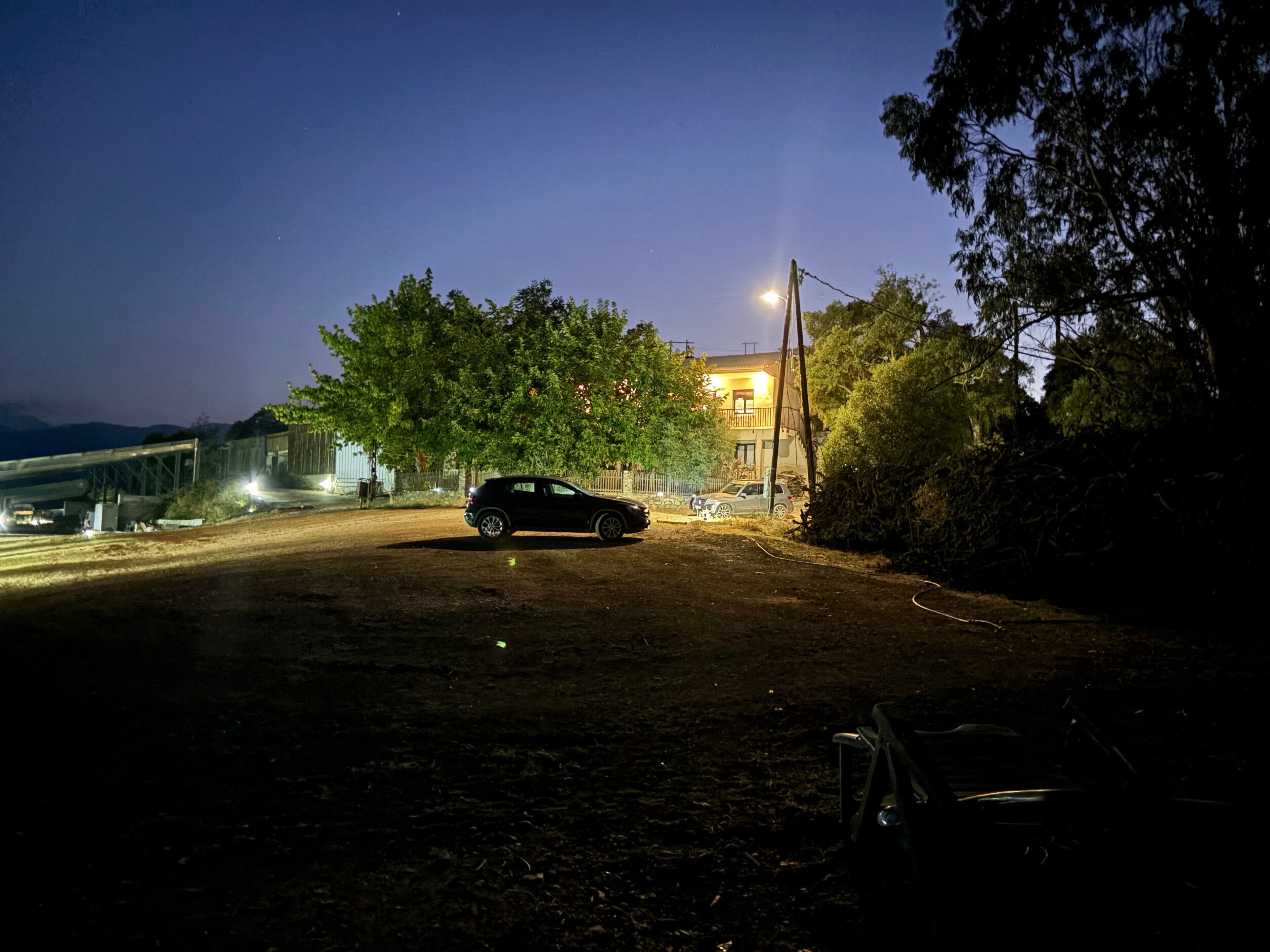 Une voiture est garée sur un chemin de terre, au crépuscule, avec un bâtiment visible au loin.