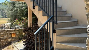 Eine Treppe mit Metallgeländer führt zu einer Zwischenebene, umgeben von einer Steinmauer und einem Garten mit Pflanzen und einem Holzstapel.