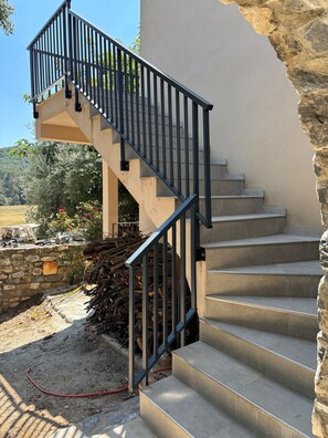 A staircase with a metal railing leading to a landing, surrounded by a stone wall and a garden with plants and a pile of firewood. - Apartment on a Farm, Near the Sea with Mountain View, Wi-Fi, and Air Conditioning (Antisanti)