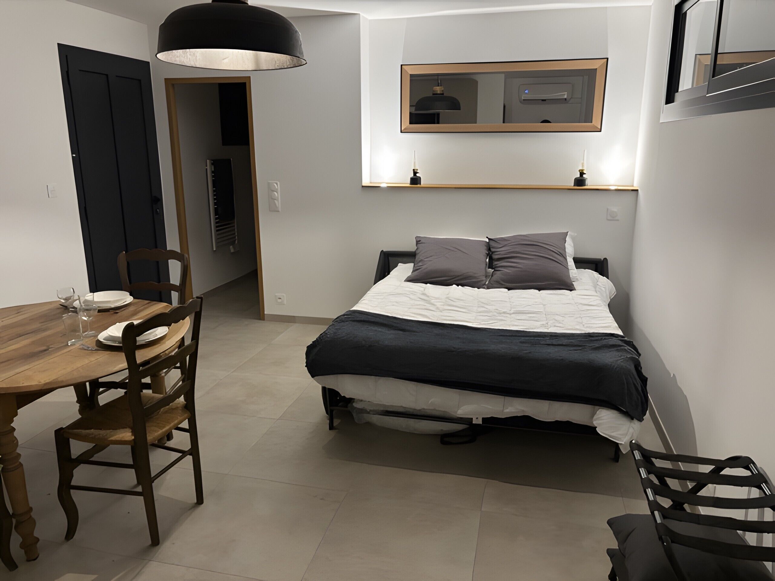 Ein Schlafzimmer mit einem Bett, einem Nachttisch und einem Sessel. Eine Tür führt in einen anderen Raum, und über dem Bett befindet sich eine kleine Ablagefläche.