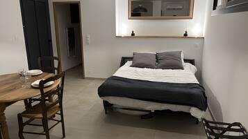 Ein Schlafzimmer mit einem Bett, einem Nachttisch und einem Sessel. Eine Tür führt in einen anderen Raum, und über dem Bett befindet sich eine kleine Ablagefläche.