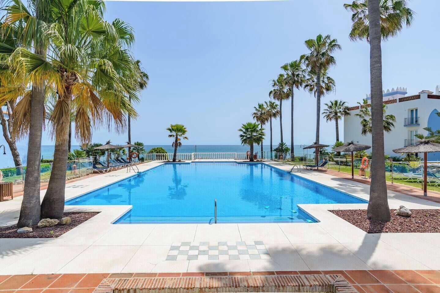 Une piscine avec vue sur la mer, entourée de palmiers et bordée d’un bâtiment doté d’un balcon.