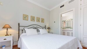 Een opgemaakt bed met witte lakens en kussens, een nachtkastje met een lamp, een wandplank, en een badkamer zichtbaar door een geopende deur.