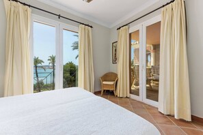 Une chambre avec un grand lit, un fauteuil en osier et une vue sur un balcon donnant sur une piscine.
