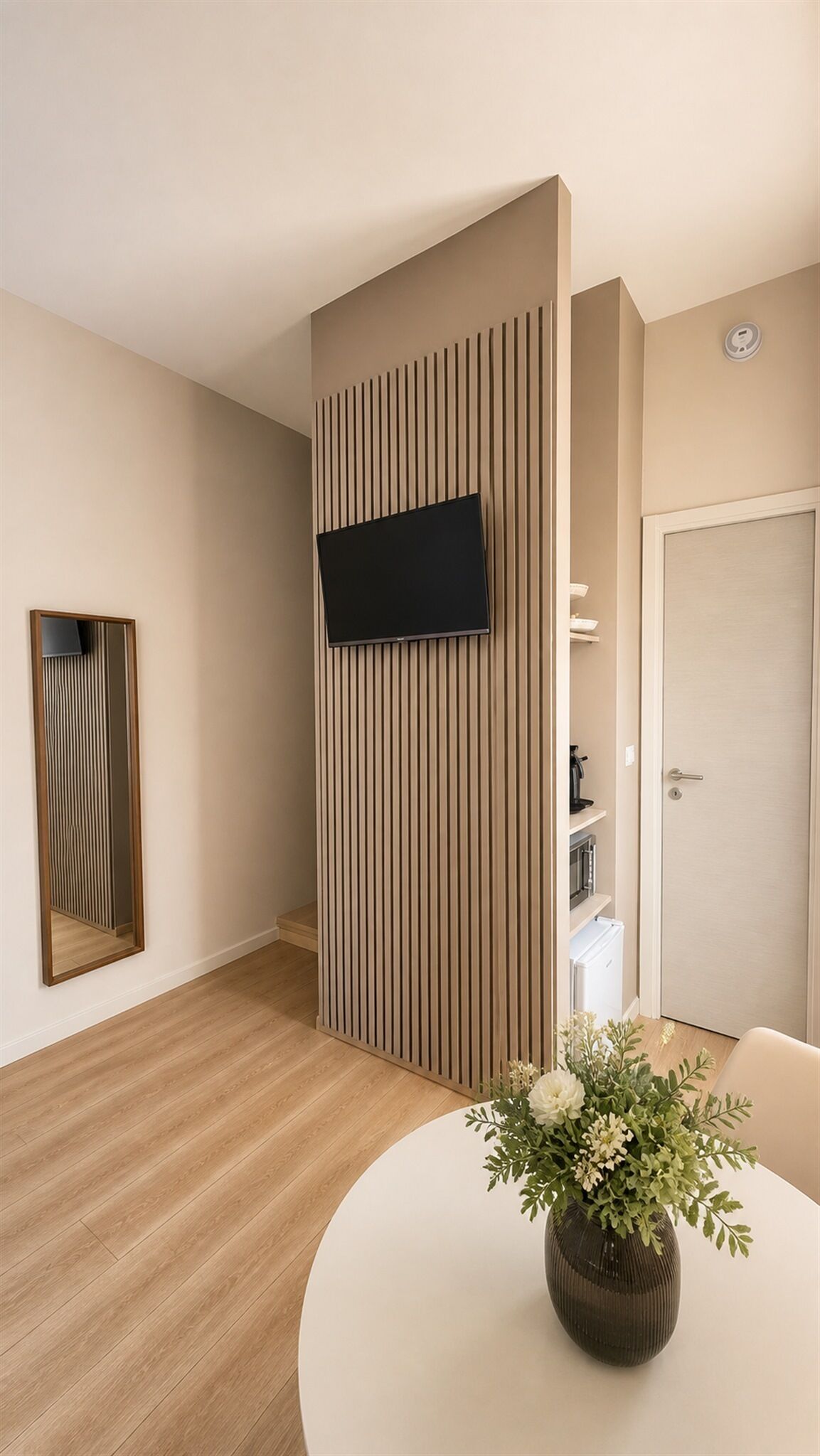 Moderni hotellihuone, jossa on seinään kiinnitetty litteänäyttöinen televisio, puupaneeliseinä, pyöreä ruokapöytä, jossa on maljakko täynnä kukkia, sekä ovi, jossa on kahva.