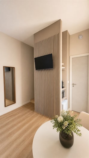 A modern hotel room with a flat-screen TV mounted on a wooden panel wall, a round dining table with a vase of flowers, and a door with a handle.
