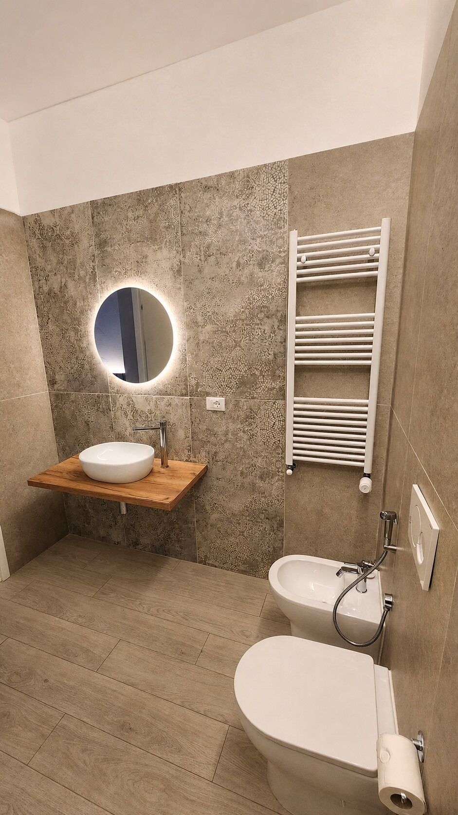 Een moderne badkamer met een houten wastafelmeubel, een ronde spiegel met verlichting, een handdoekrek, een toilet en een bidet.