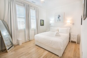 A hotel room with a large bed, a bedside table, a nightstand, a mirror, and a window with blinds. - Walk to King St | Spacious Charleston Stay (Charleston)