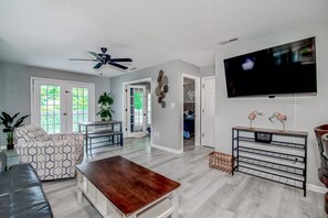 Ein modernes Wohnzimmer mit Sofa, Couchtisch und Fernseher. Es gibt einen Deckenventilator, einen Essbereich und einen Flur, der in einen anderen Raum führt.