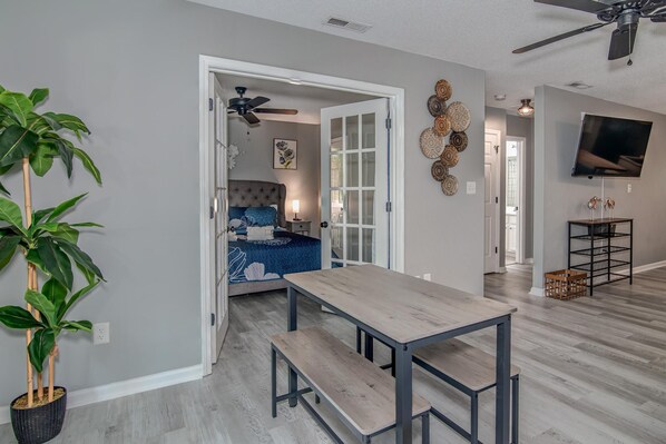 A modern living room with a dining area, a large window, and a ceiling fan. - Hammock Haven | Walk to Marshwalk! (Murrells Inlet)