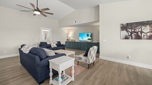 A modern living room with a dark blue sofa, a white coffee table, and a patterned armchair. A ceiling fan and a flat-screen TV are visible in the background. - Mollymawk Cottage: Peaceful Pool Home Near Beaches, Golf & Island Adventures (Sanibel)