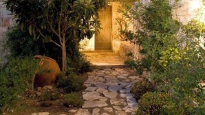 A stone pathway leading to a wooden door, surrounded by lush greenery and a stone building with a tiled roof. - Aphrodite's Oasis with Private Pool (Pomos)