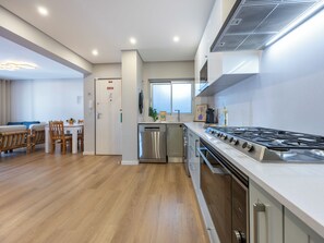 A modern kitchen with a stainless steel stove, a dishwasher, and a dining area with wooden chairs and a white table. - Algarve Sun Apartment - Family Home in Portimão (Portimão)