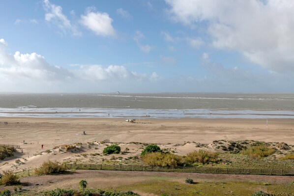 A wide sandy beach with a fence in the foreground, a clear sky, and a distant view of the sea.
