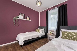 A room with two beds, a small table, a hanging plant, and a window with curtains.