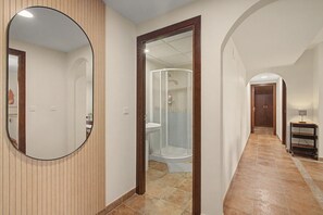 Un pasillo con pared de paneles de madera, un espejo, cabina de ducha con mampara de vidrio y suelo de baldosas.