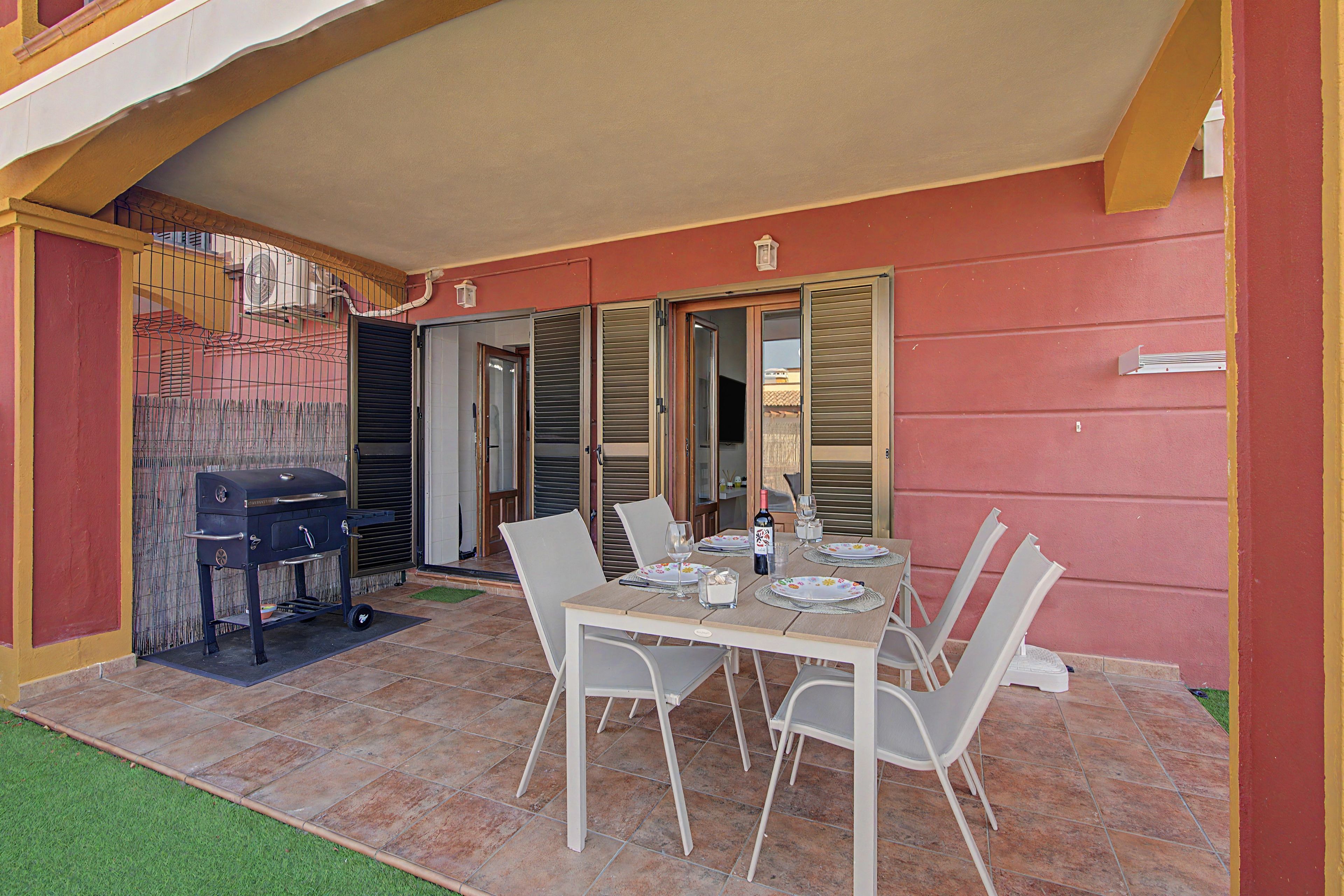 Eine Außenpatio mit einem Esstisch, gedeckt für vier Personen, umgeben von Stühlen, einem Grill und einem überdachten Bereich mit einer Tür.
