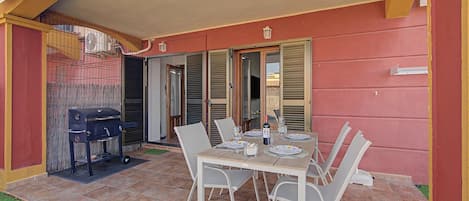An outdoor patio with a dining table set for four, surrounded by chairs, a barbecue grill, and a covered area with a door.