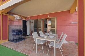 An outdoor patio with a dining table set for four, surrounded by chairs, a barbecue grill, and a covered area with a door.