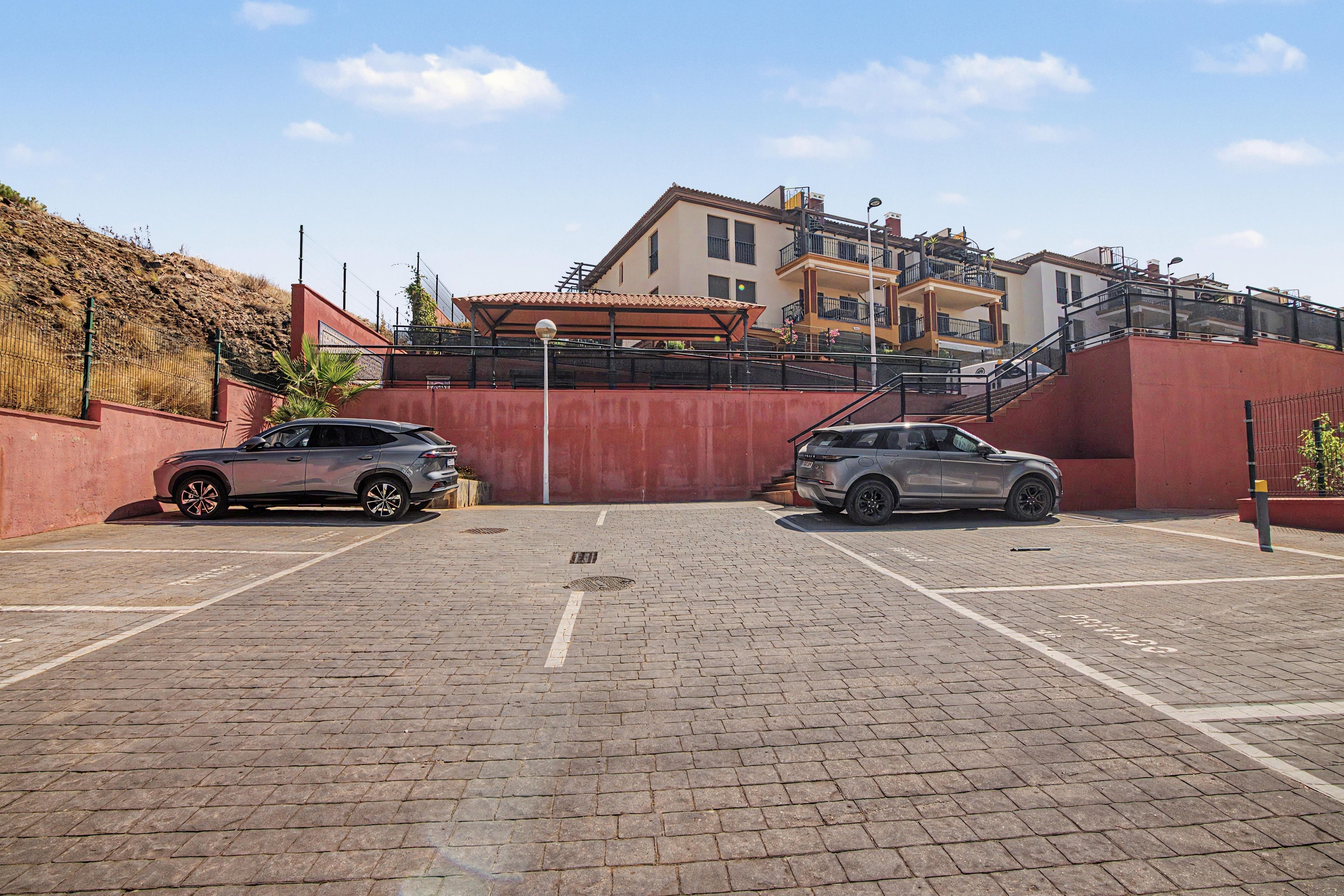 Ein Parkplatz mit zwei Autos, einer roten Wand und einem Gebäude mit mehreren Etagen und Balkonen.