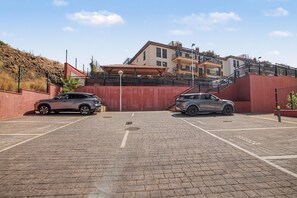 A parking area with two cars, a red wall, and a building with multiple floors and balconies.