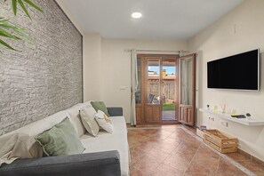 A modern living room with a stone-textured wall, a grey sofa with cushions, a wooden door leading to a balcony, and a mounted TV. - Brisa of Guadiana – Ayamonte Holiday Home by the River, Pool, Bikes, Pet Friendly (Ayamonte)