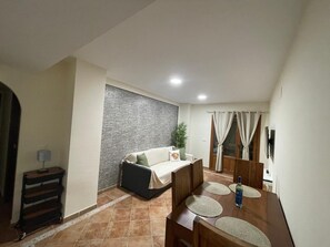 A living room with a sofa, a dining table with chairs, a TV, and a wall-mounted light fixture. - Brisa of Guadiana – Ayamonte Holiday Home by the River, Pool, Bikes, Pet Friendly (Ayamonte)