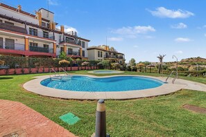 Una piscina con una terraza circundante, una zona verde y un edificio con balcones al fondo.
