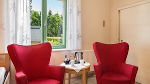 Een kamer met twee rode stoelen, een tafeltje met een waterkoker en een kop, en een raam met groene kaders en witte gordijnen.
