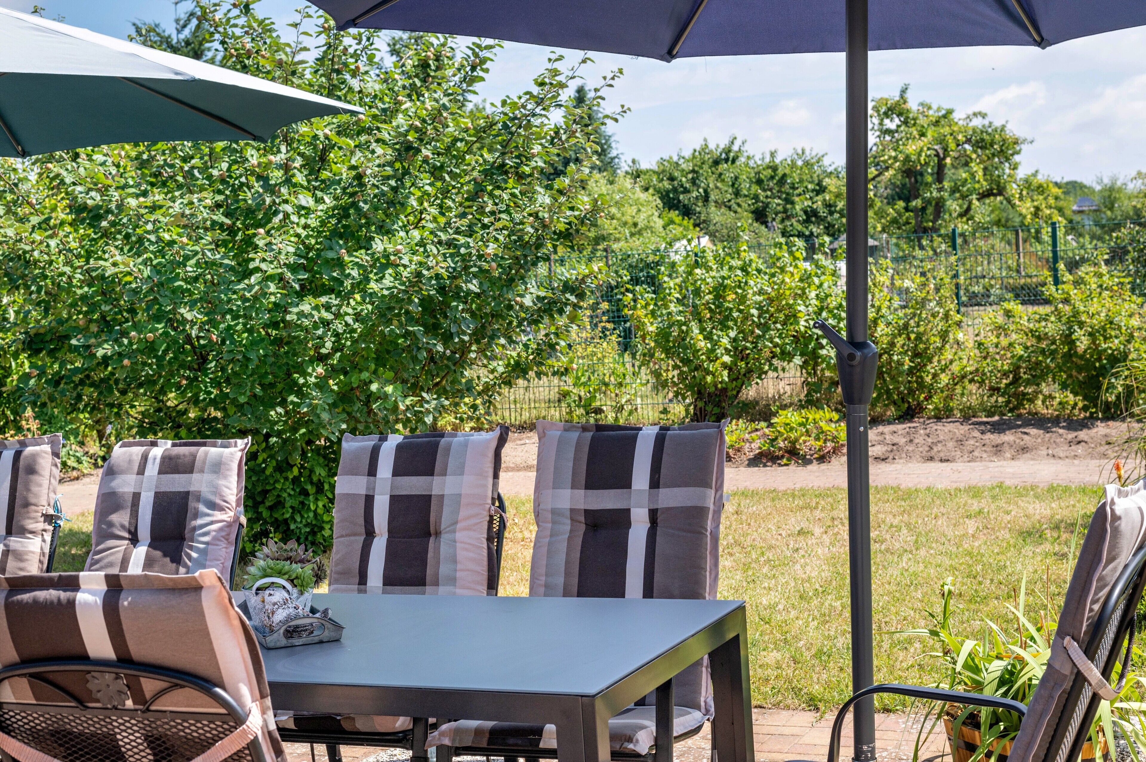 Eine Terrasse im Freien mit Tisch und Stühlen, umgeben von viel Grün und einem Zaun.