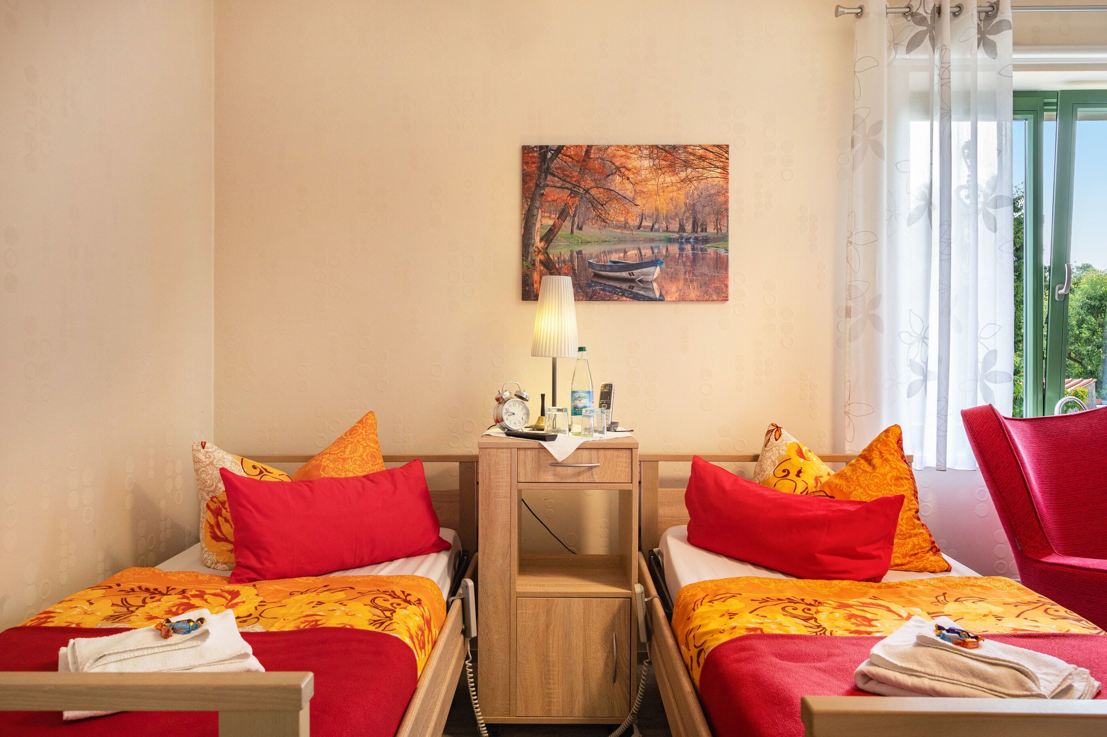 Ein Hotelzimmer mit zwei Betten, einem Nachttisch, einer Lampe, einem Gemälde und Blick ins Freie.