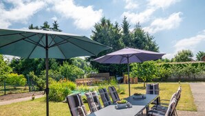 Een terras buiten met een tafel, stoelen, twee parasols en een tuin op de achtergrond.