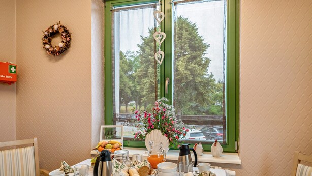 Een ontbijttafel gedekt met borden, kopjes, een broodmand, een bloevas en uitzicht op een groen raam met een krans.