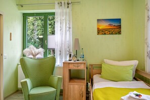 Une pièce comprenant un lit, une chaise, une table de chevet avec une lampe, un tableau au mur et une fenêtre avec des rideaux.