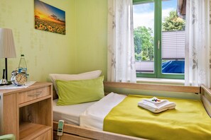 Un dormitorio con una cama individual, una mesita de noche con lámpara y reloj, una ventana con cortinas y un cuadro en la pared.